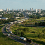 Vista aérea de Campo Grande com rodovia e veículos de carga representando a integração comercial da cidade pela Rota Bioceânica