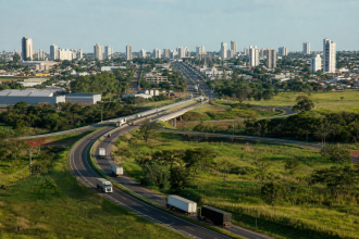 Vista aérea de Campo Grande com rodovia e veículos de carga representando a integração comercial da cidade pela Rota Bioceânica