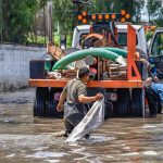 Trabalhadores caminham por uma área alagada próxima a um caminhão durante uma operação urbana após chuva intensa.