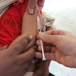 Profissional de saúde aplica uma vacina em uma criança dentro de uma unidade de atendimento.