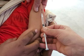 Profissional de saúde aplica uma vacina em uma criança dentro de uma unidade de atendimento.