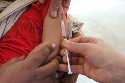 Profissional de saúde aplica uma vacina em uma criança dentro de uma unidade de atendimento.