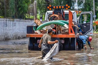 Trabalhadores caminham por uma área alagada próxima a um caminhão durante uma operação urbana após chuva intensa.