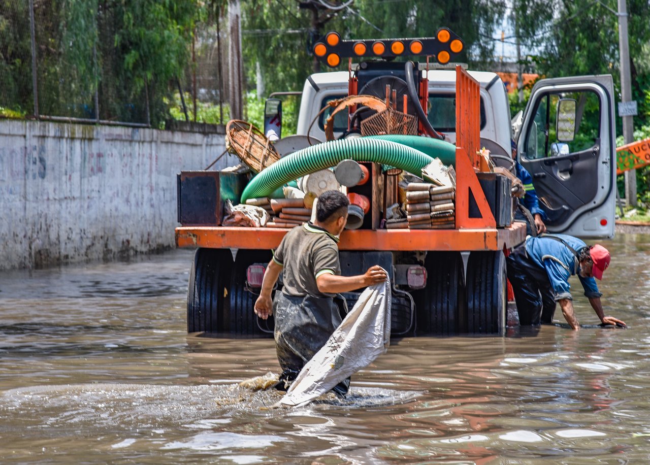 Trabalhadores caminham por uma área alagada próxima a um caminhão durante uma operação urbana após chuva intensa.